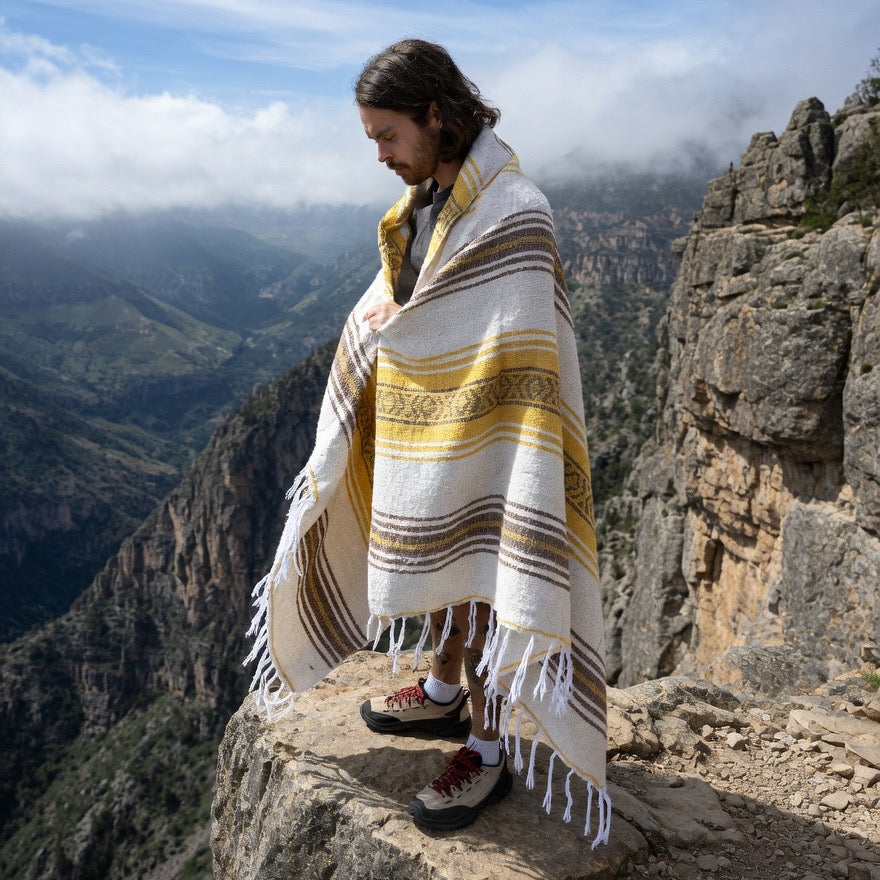 Person wrapped in a striped blanket standing on a rocky mountain ledge with a scenic view.