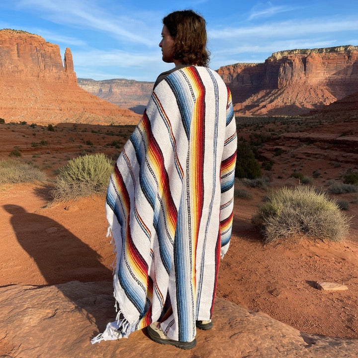 Person wrapped in a colorful striped blanket standing in a desert landscape with red rock formations.