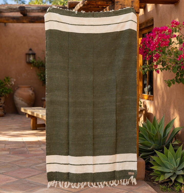Green and beige blanket hanging on a wooden rack outdoors with plants and a clear sky.