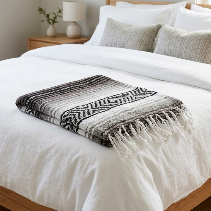 Bed with white bedding and a black and white patterned throw blanket, nightstand with lamp and plant in the background.