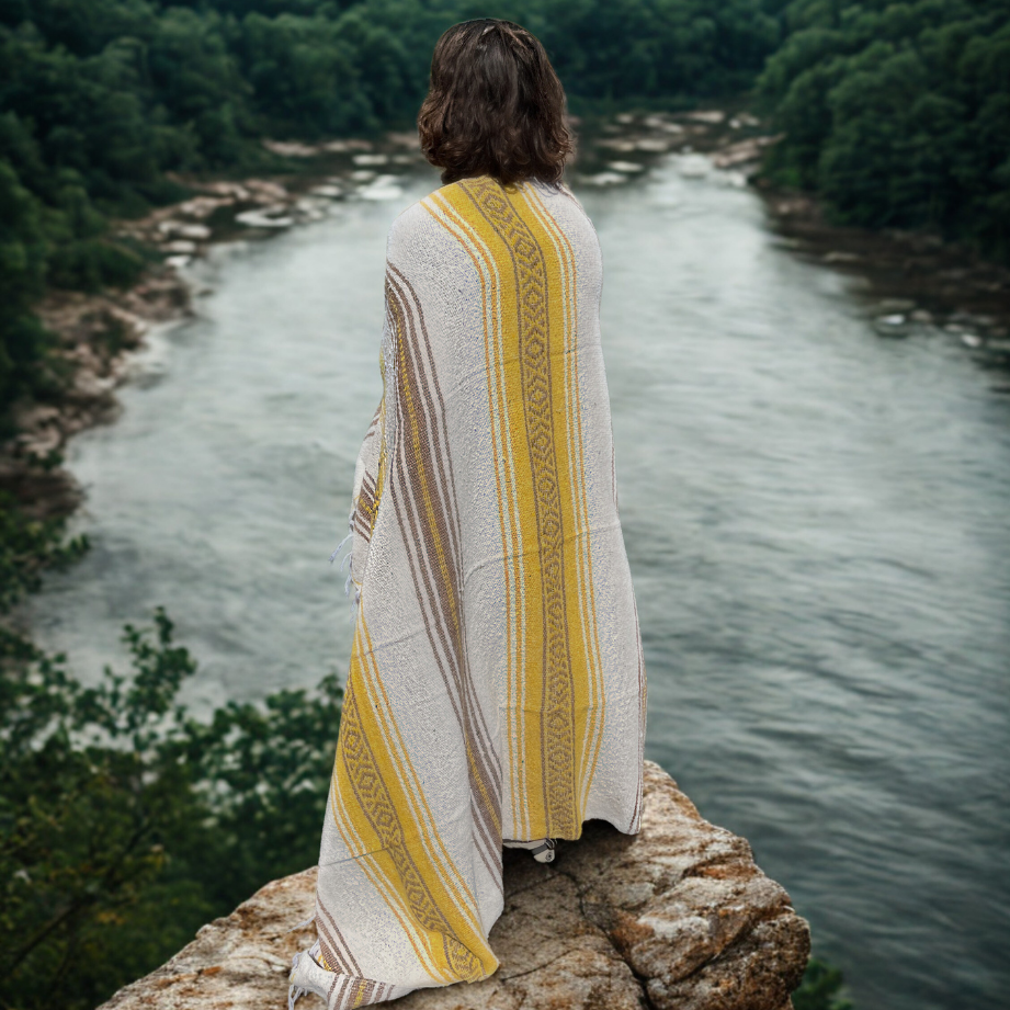 Person wrapped in a yellow and white patterned blanket overlooking a river with trees.