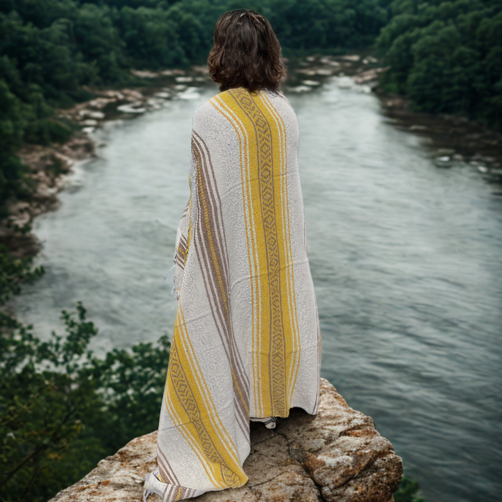Person wrapped in a yellow and white patterned blanket overlooking a river with trees.