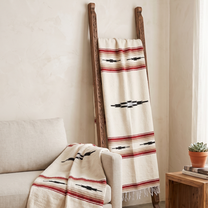 A cream woven throw blanket with red and black Southwestern-style stripes and fringe, draped over a light beige sofa and hanging on a wooden ladder in a styled living room.