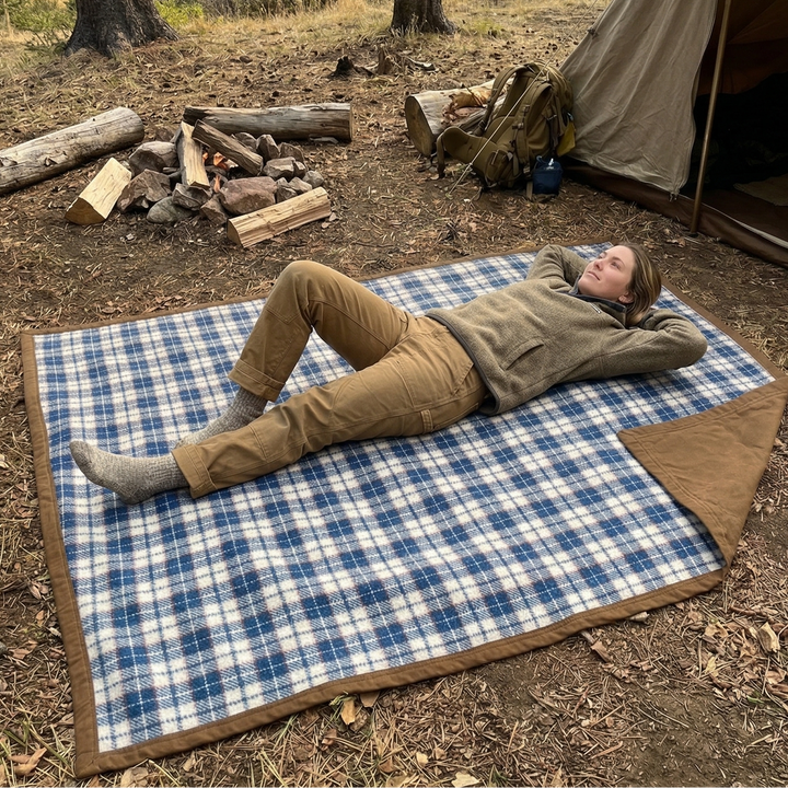 Person lying on a blue plaid blanket in front of a tent by a lake.