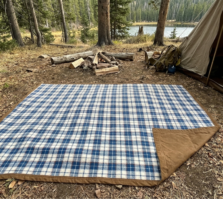 Blue and white checkered blanket on the ground next to a tent in a forest setting.